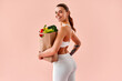 © HBS - A fit young woman is holding a bag of vegetables against a soft pastel background in a studio. Concept of healthy eating and fitness.
