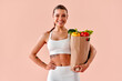 © HBS - A beautiful, smiling, athletic girl in white fitness clothing is holding a bag of vegetables and smiling against a light pink background. Healthy eating and fitness.