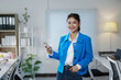 ©  NCST Studio - Smiling businesswoman enjoying a break while holding her smartphone, standing confidently in a bright, modern office workspace