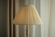 © Thedinindu - A beige pleated lampshade sits atop a cream-colored lamp base, illuminating a softly lit room.