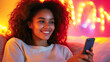 © Bonsales - Smiling young woman with curly hair using mobile phone in her bedroom decorated with string lights