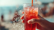 © britaseifert - Close-up of hands holding a cold drink with a straw, condensation on the glass, beach or pool blurred in the background