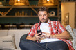 © Volodymyr - Man looking at phone at home, browsing mobile apps, reading news, chatting or shopping online, holding smartphone. Attractive man in casual clothes at the living room, talking on mobile phone.