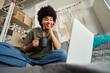© insta_photos - Happy gen z African American girl using laptop sitting at home bed drinking coffee. Young smiling Afro woman looking at computer relaxing with cup of tea in hands watching online tv series. Low angle.
