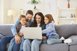 © Studio Romantic - Smiling parents sitting on sofa with two children, hugging, using laptop together. Happy cheerful family watching something fun on screen, enjoying quality time and having fun in living room at home.