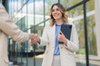 © Dexon Dee - Confident businesswoman smiling and shaking hands with a colleague outdoors in front of a modern glass building. Friendly greeting symbolizing collaboration and trust