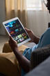 © pressmaster - Vertical medium shot of African American man sitting on couch at home, watching movie or TV show trailer on streaming app on tablet