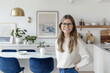 © Katerina Bond - Smiling young female interior designer with long hair and glasses standing in bright modern kitchen with minimalist decor and blue velvet chairs, natural light.