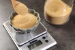 © New Africa - Putting brown sugar into bowl on digital kitchen scale at grey table, closeup