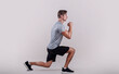 © Prostock-studio - Side view of young fit guy in sportswear doing lunge on light studio background. Professional millennial bodybuilder warming out before strength training, doing aerobic exercises