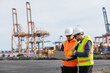 © kamonrat - Two engineers wearing safety vests and helmets stand at a shipping port, discussing logistics while using a laptop. Large cranes and stacked cargo containers are visible in the background.
