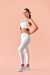© HBS - A vertical image of a sporty woman in white fitness clothing posing against a soft peach background in a studio. Fitness training, yoga, pilates, stretching.