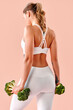 © HBS - A sporty girl in white workout clothing stands with her back to the camera, holding fresh broccoli like dumbbells. Vertical image with the girl set against a soft pink background.
