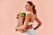 © HBS - A sporty girl holds a grocery bag filled with fresh vegetables and smiles against a soft peach background. A concept of healthy eating and fitness — caring for health and the body.