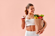 © HBS - Smiling girl in sportswear holds a creamy dessert in one hand and a bag of vegetables and healthy food in the other, smiling as she looks at the bag.The balance between temptation and healthy eating.