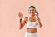 © HBS - A happy young woman in a white sports bra and leggings is dancing in blue headphones against a soft peach background in a studio. Living a sporty lifestyle.