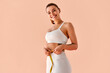 © HBS - Smiling fit young woman in white sportswear measuring her waist with a tape measure on a soft peach background. Concept of healthy lifestyle, fitness tracking, and workout motivation.