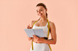 © HBS - A confident, attractive woman — a fitness trainer — standing against a soft peach background, holding and writing in a notebook with a nutrition and workout plan. Personal trainer.