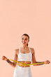 © HBS - Vertical image of a fit young woman in a white sports bra and leggings posing with a measuring tape against a soft peach background. Copy space.