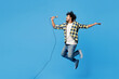 © ViDi Studio - Full body young happy Indian man wear yellow checkered shirt casual clothes jump high sing song in microphone at karaoke club isolated on plain blue cyan background studio portrait. Lifestyle concept.