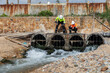 © kamonrat - Two environmental engineers inspect industrial drainage pipes with flowing wastewater, assessing water quality and potential pollution risks near a facility with rusted infrastructure.