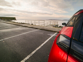Wall Mural - Red car parked in car park with stunning view on nature scenery with ocean and mountains and cloudy sky. Silverstrand area, Galway city, Ireland. Rent motor for a tourist trip concept.
