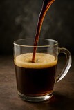 Dark coffee streams into a clear glass mug, creating a rich, frothy surface with rising steam on a dark background