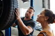© peopleimages.com - Customer, tire and mechanic in garage for maintenance, inspection and car service assessment. Woman, wheel and check in auto repair workshop for motor performance, quality assurance or vehicle safety