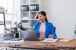 © David - A focused asian woman accountant analyzing paperwork while calculating expenses using laptop and calculator at her office desk.