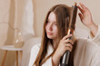 © st.kolesnikov - Girl styling long hair with curler tool, creating smooth waves in cozy home setting.