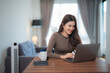 © Tj - Happy freelancer working remotely from home sitting at a wooden table with a laptop, a smartphone and a cup of coffee, enjoying her smart working job