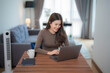 © Tj - Happy young Asian businesswoman working remotely using laptop computer while sitting at wooden table in living room with a cup of coffee and smartphone near her