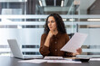 © Liubomir - A focused businesswoman is reviewing documents and working on a laptop in a modern office setting. She appears thoughtful.