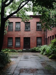 Poster - A brick building's courtyard entrance, framed by lush green foliage and a tree, shows a dark wooden door and multiple windows reflecting a cloudy sky. : Generative AI