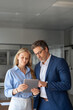 © Stock 4 You - Mature senior leader man in suit and young employee woman working together on project on tablet pad device in office. Team of two colleagues of professional business people using computer. Vertical