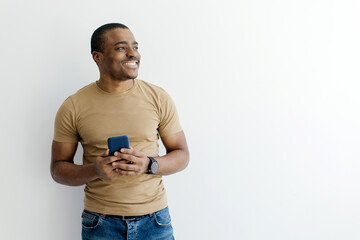 Smiling African American man holding smartphone while looking aside at copy space, handsome young black male standing in casual and relaxed posture on white studio background