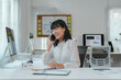 © crizzystudio - Cheerful young asian office employee sitting at a desk, engaging in a conversation on a smartphone while reviewing notes, surrounded by a computer and various documents