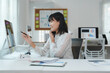 © crizzystudio - Smiling businesswoman engaging in a phone call while pointing at a computer screen, analyzing marketing data and working diligently at her office desk in a modern workspace