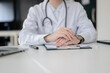 © bongkarn - Doctor with stethoscope clasping hands on clipboard with document sitting at table in diagnosis room