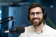 © Tetiana - Close-up portrait of a young smiling man sitting in the office wearing headphones and earmuffs in front of a desk with a microphone and looking at the camera
