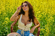 © czamfir - Portrait of a beautiful woman with long hair in a blouse and jeans sitting on a chair in a field of blooming rapeseed