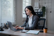 © MINAE - Young Asian businesswoman sitting at desk and working with laptop computer in modern office, focused entrepreneur typing on keyboard, managing business tasks and projects