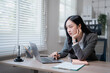 © MINAE - Pensive Asian businesswoman working on a laptop at a modern office, resting her chin on her hand while contemplating a project at her desk by the window