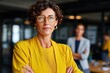 © olgasun - Confident businesswoman in yellow blazer with glasses stands in modern office, with her team members visible in the blurry background, looking directly at the camera.
