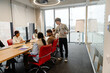 © Drobot Dean - A young White man with glasses is leading a team discussion during an internship session with a multinational group of people in a modern office setting with a large wooden table and large windows.