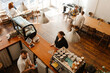 © Drobot Dean - A barista interacts with a woman at the counter of a cozy cafe. The cafe features wooden furniture, a modern espresso machine, and several people sitting at tables enjoying drinks and conversation.