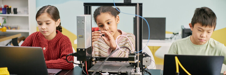 Website header shot of children of diverse backgrounds collaborating on a technology project in modern classroom environment. They are working with electronic devices and showing focused concentration