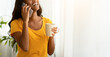 © Prostock-studio - Charming black woman speaking on cellphone with cup of aromatic coffee at home. Attractive African American lady having conversation on mobile phone near window indoors