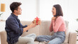 © Prostock-studio - Korean Boyfriend Giving Wrapped Gift Box To Surprised Girlfriend Celebrating Christmas Holiday Or Valentine's Day Together Sitting On Sofa At Home. Romance, Holidays Pressents And Celebration
