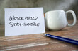 © Maria Marganingsih - Business motivational quotes - Work hard, stay humble. Text messages on a white paper with cup of coffee or tea and a pen on the wooden table. Positive slogan motivation concept.